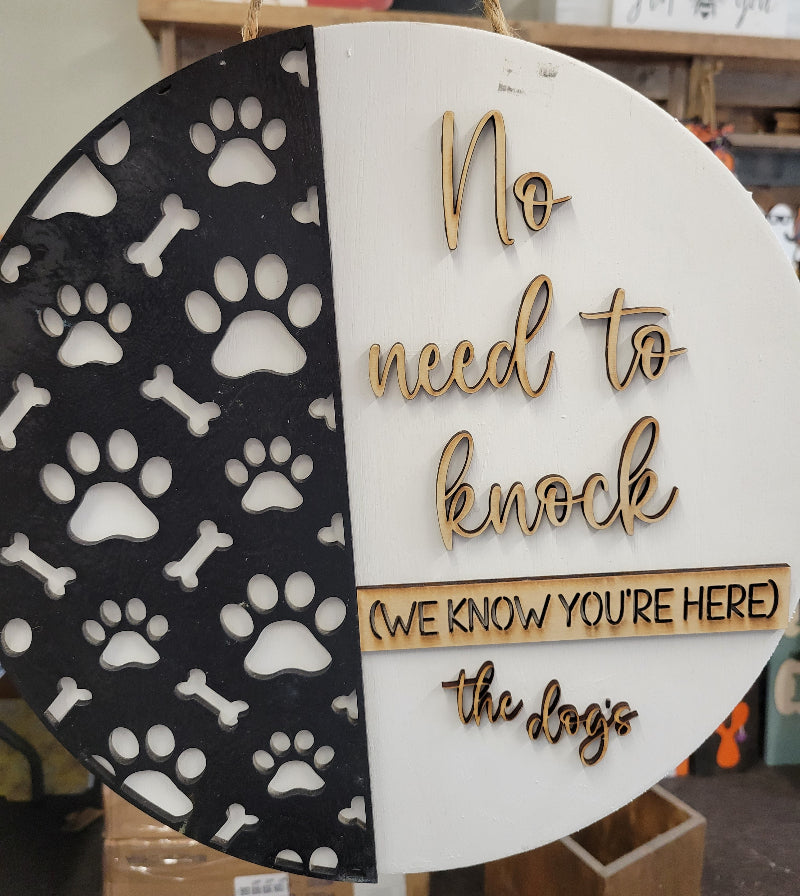 A round doorhanger with a white background featuring black paw prints and text that reads 'NO NEED TO KNOCK HERE' along with 'THE DOGE'S' logo.
