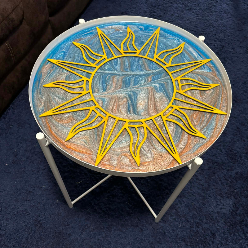 A round glass-top side table with a white metal frame, featuring a large yellow sun design over a marbled blue, white, and orange artwork on the tabletop, set on a dark blue carpet.