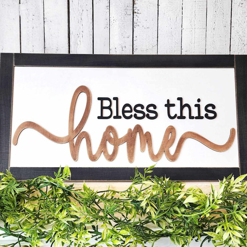 A DIY craft kit sign with the words 'BLESS THIS HOME' in a cursive font, mounted on a black frame with a wooden border, displayed against a backdrop of greenery.
