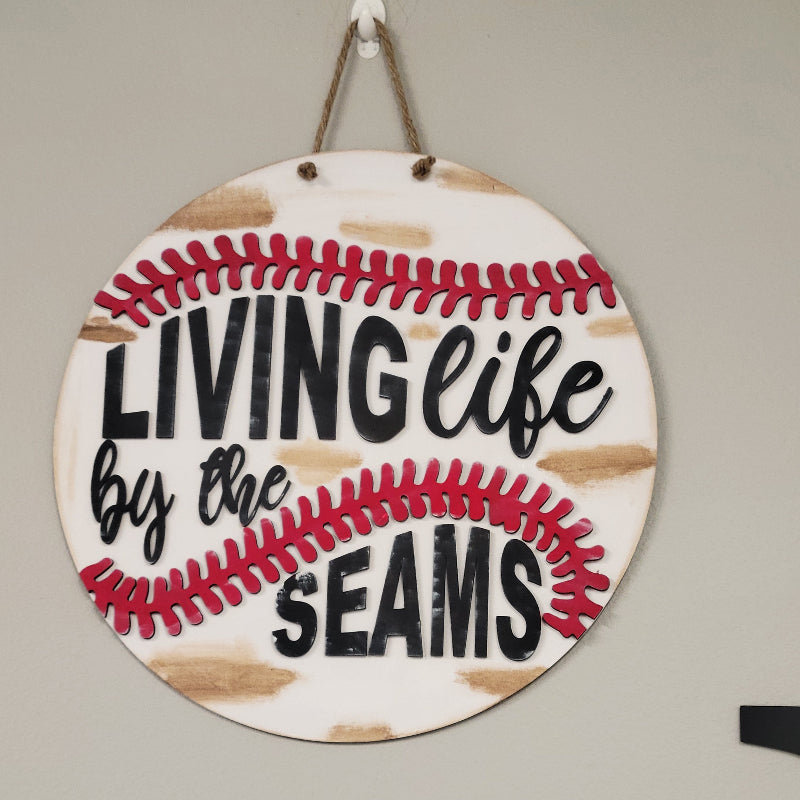 Round wooden door hanger with a baseball design and the text 'LIVING life by the SEAMS' in red and black.