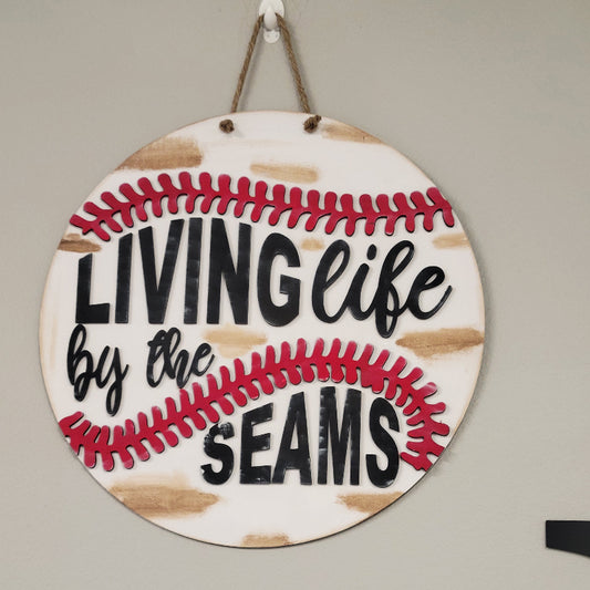 Round wooden door hanger with a baseball design and the text 'LIVING life by the SEAMS' in red and black.