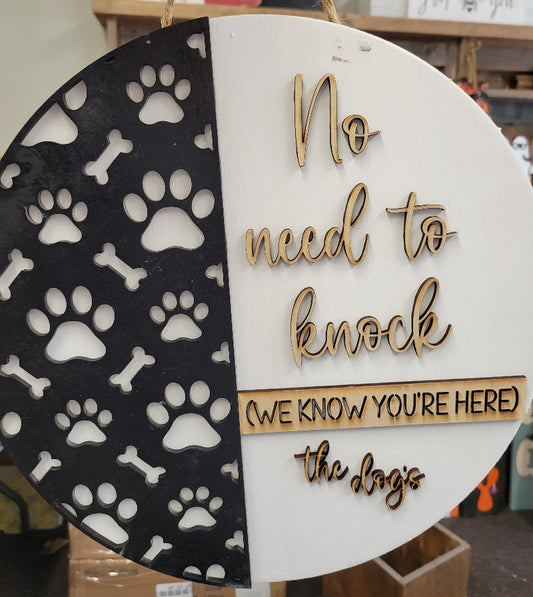 A round doorhanger with a white background featuring black paw prints and text that reads 'NO NEED TO KNOCK HERE' along with 'THE DOGE'S' logo.