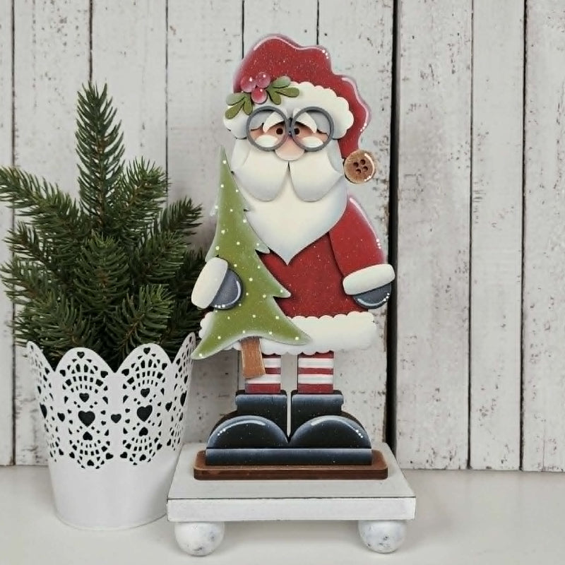 Decorative Santa Claus figure holding a green tree on a white surface with a wooden panel background.