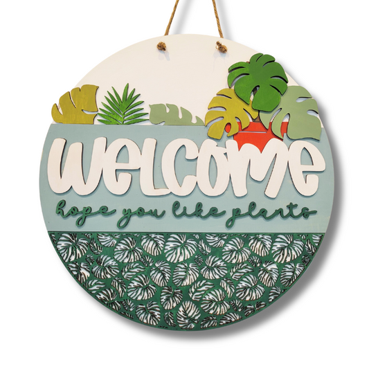 Round wooden sign with tropical leaf designs and 'Welcome hope you like plants' text on a white background