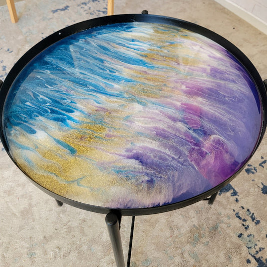 A round black metal side table with a glossy fluid art resin top featuring swirls of blue, purple, gold, and white, shown in a room setting.