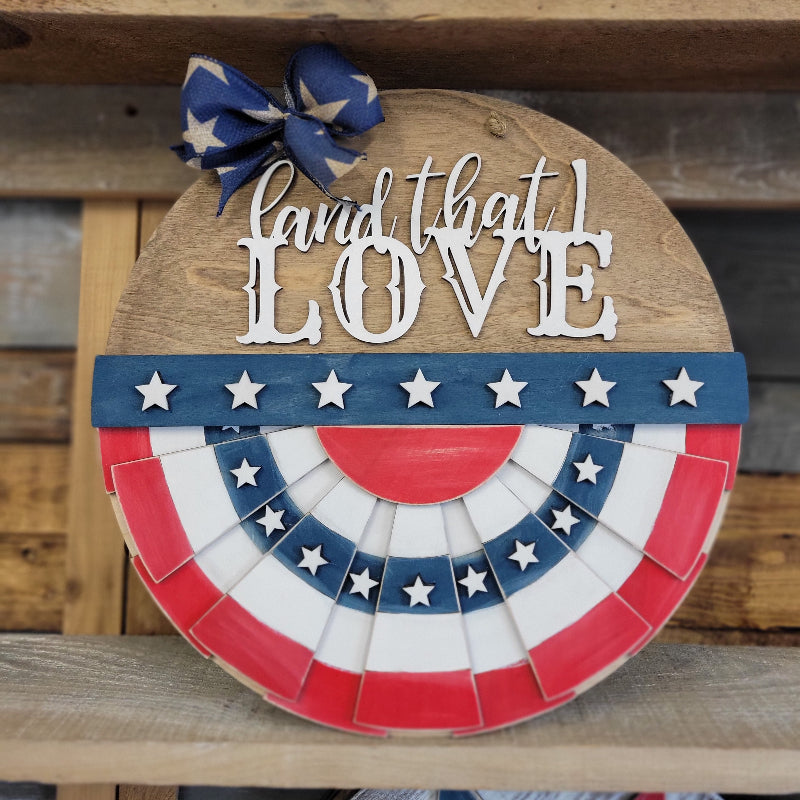 A round doorhanger featuring a patriotic design with a white banner that says 'Land that I love' and a blue bow, red and white stars, and a wooden background.
