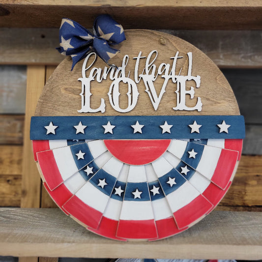 A round doorhanger featuring a patriotic design with a white banner that says 'Land that I love' and a blue bow, red and white stars, and a wooden background.
