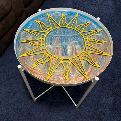 A round glass-top side table with a white metal frame, featuring a large yellow sun design over a marbled blue, white, and orange artwork on the tabletop, set on a dark blue carpet.