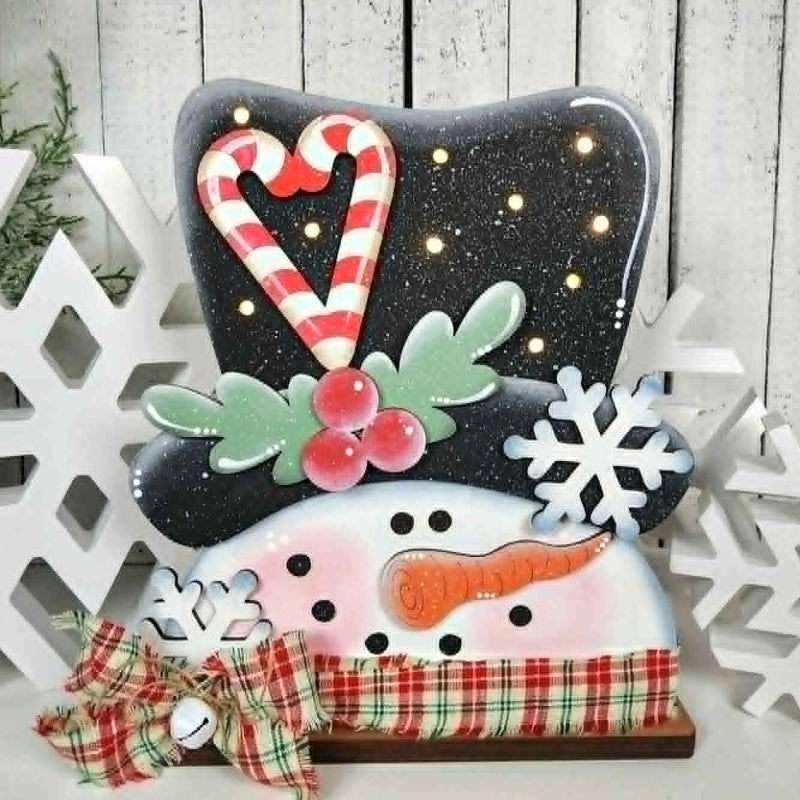 Decorative snowman figure with candy cane hat and plaid scarf, surrounded by white snowflakes on a wooden background.