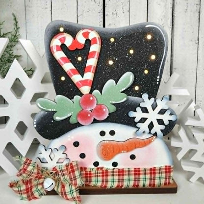 Decorative snowman figure with candy cane hat and plaid scarf, surrounded by white snowflakes on a wooden background.