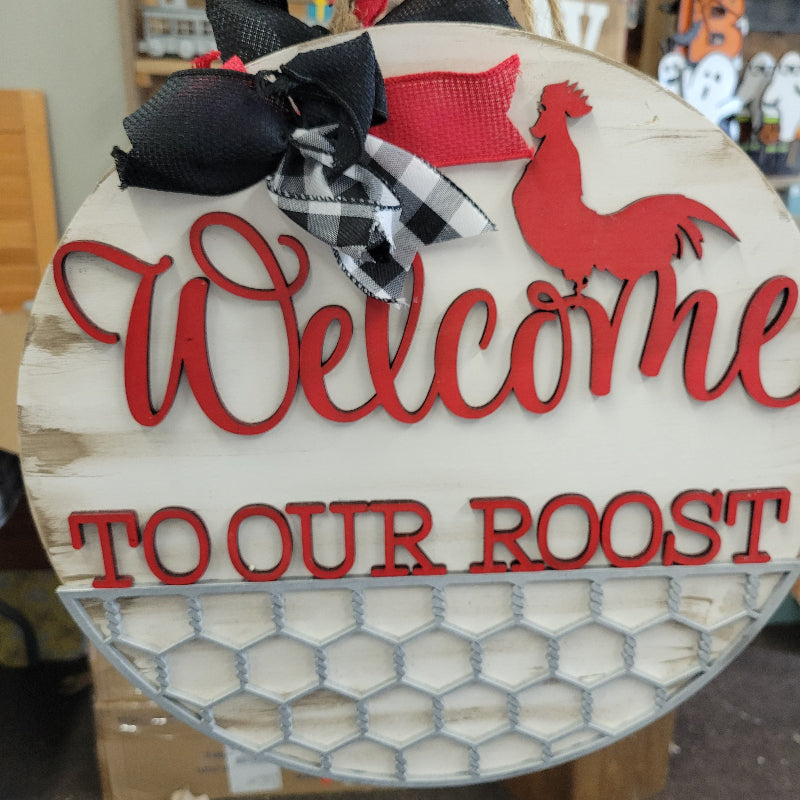 A round wooden doorhanger with a white background, red text that reads 'Welcome to our ROOST' and a red rooster graphic. It is adorned with a black and red bow at the top.