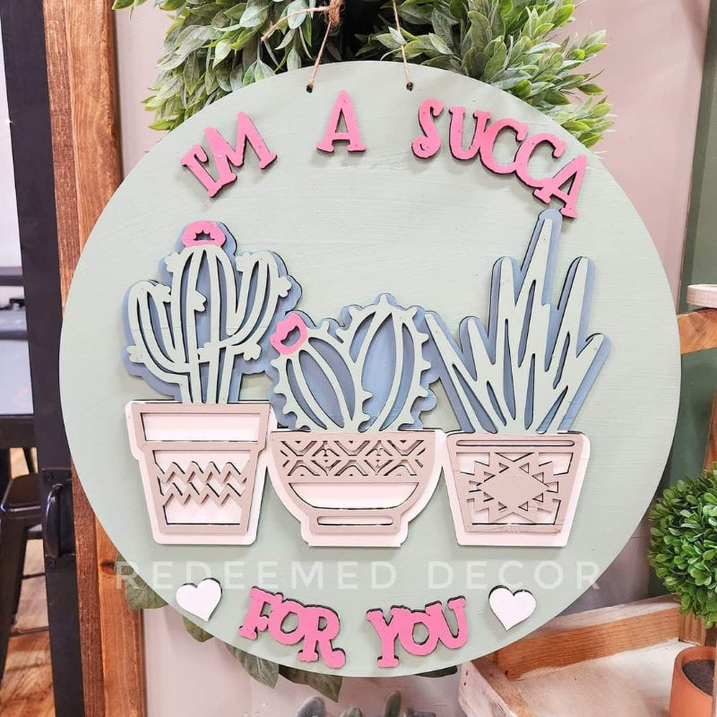A round doorhanger with a green background featuring text and illustrations of succulent plants in terracotta pots, with a punny phrase 'I'm a Succa for You'.