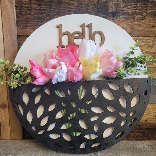 A round black decorative tray with a floral arrangement in it, placed against a wooden background. The tray has a pocket for seasonal decoration, and text that reads 'HELLO'.